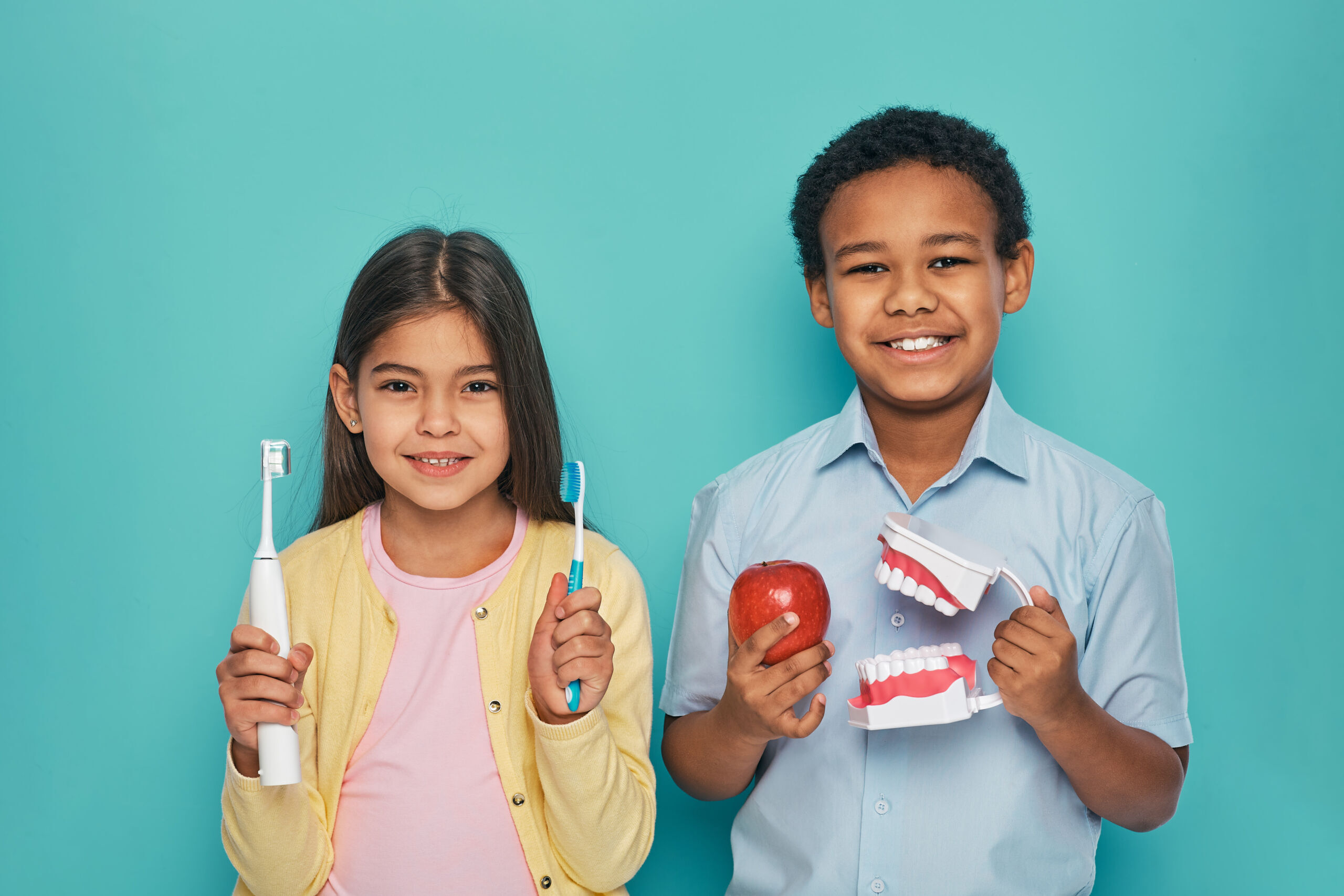 Asian girl and African American boy advertising oral hygiene and brushes teeth. Kids holding manual and electric ultrasound toothbrushes Two children smiling while holding dental care items including a toothbrush, electric toothbrush, and apple against a turquo.