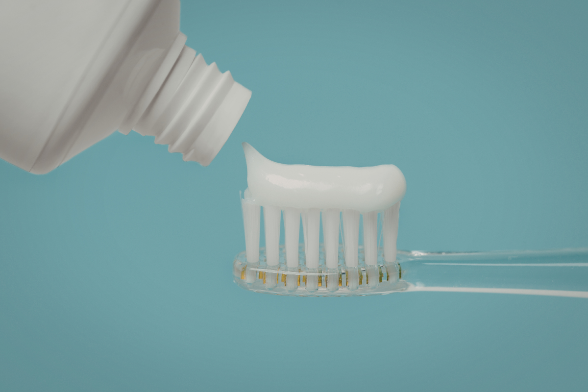Natural Toothpaste vs Fluoride Toothpaste Whats Safer Toothpaste being dispensed onto a toothbrush against a turquoise background.